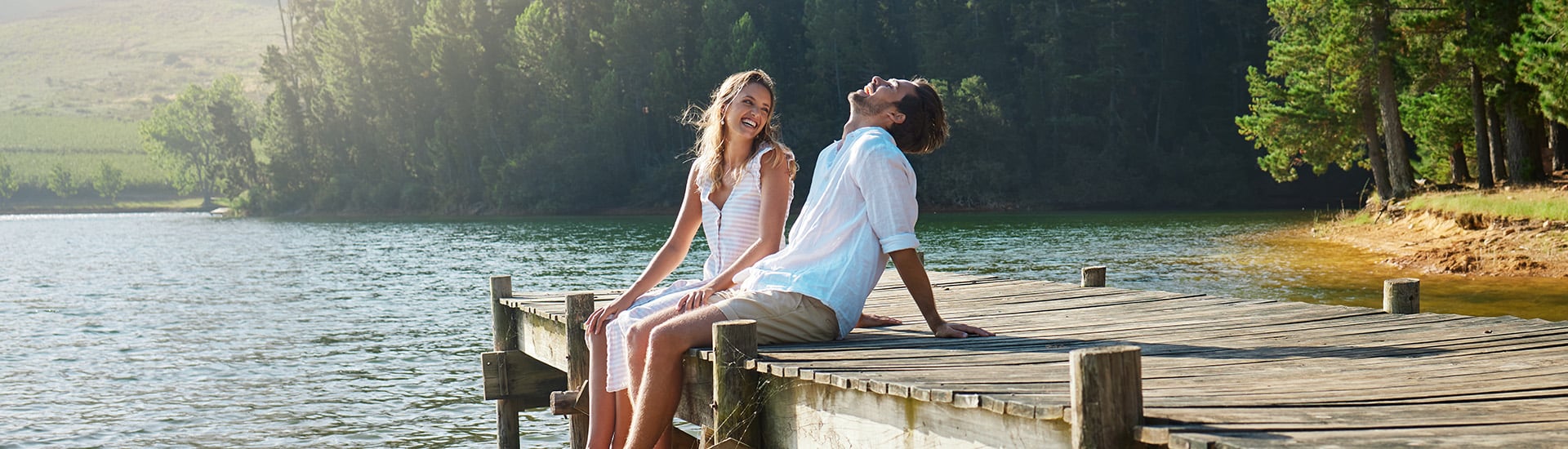 Pärchen sitzt auf einem Steg am See in der Frühlingssonne und lacht - genießt den Urlaub mit einer Reiseversicherung