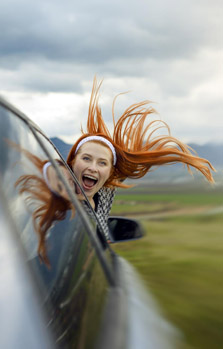 Rothaarige Frau schaut aufgeregt und glücklich aus dem Seitenfenster eines Autos, gut versichert auf alle Reisen