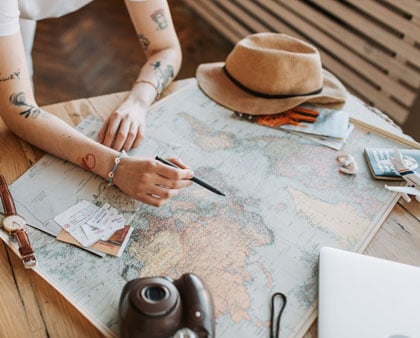 Reiseplanung, Hände die etwas auf einer Karte markieren, Reisepass, Sonnenhut und Kamera