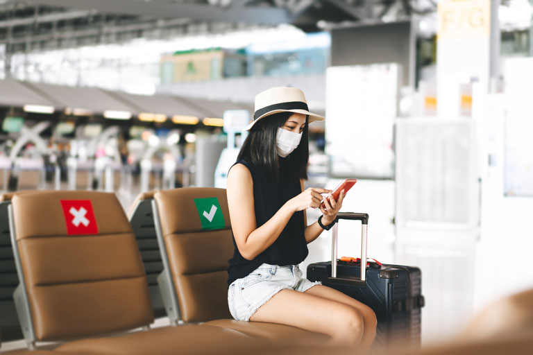 Frau am Flughafen, mit Corona Schutzmaske, tippt in ihr Telefon