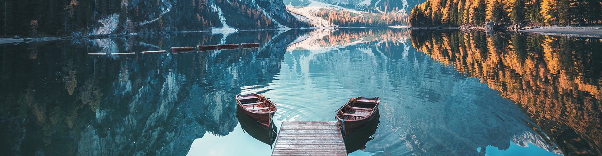 Herbstliche Stimmung am See, umgeben von Bergen, ein Steg ragt ins Wasser und zwei Boote sind davor