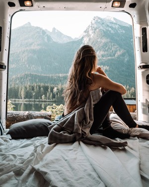 Frau mit langen Haaren, sitzt in einem Camper und genießt den Blick auf einen See