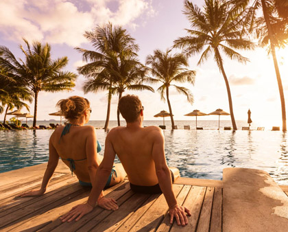 Ein Paar im Sommerurlaub, am Pool sitzend, Palmen im Hintergrund - genießt den Urlaub entspannt mit einer Reiseversicherung