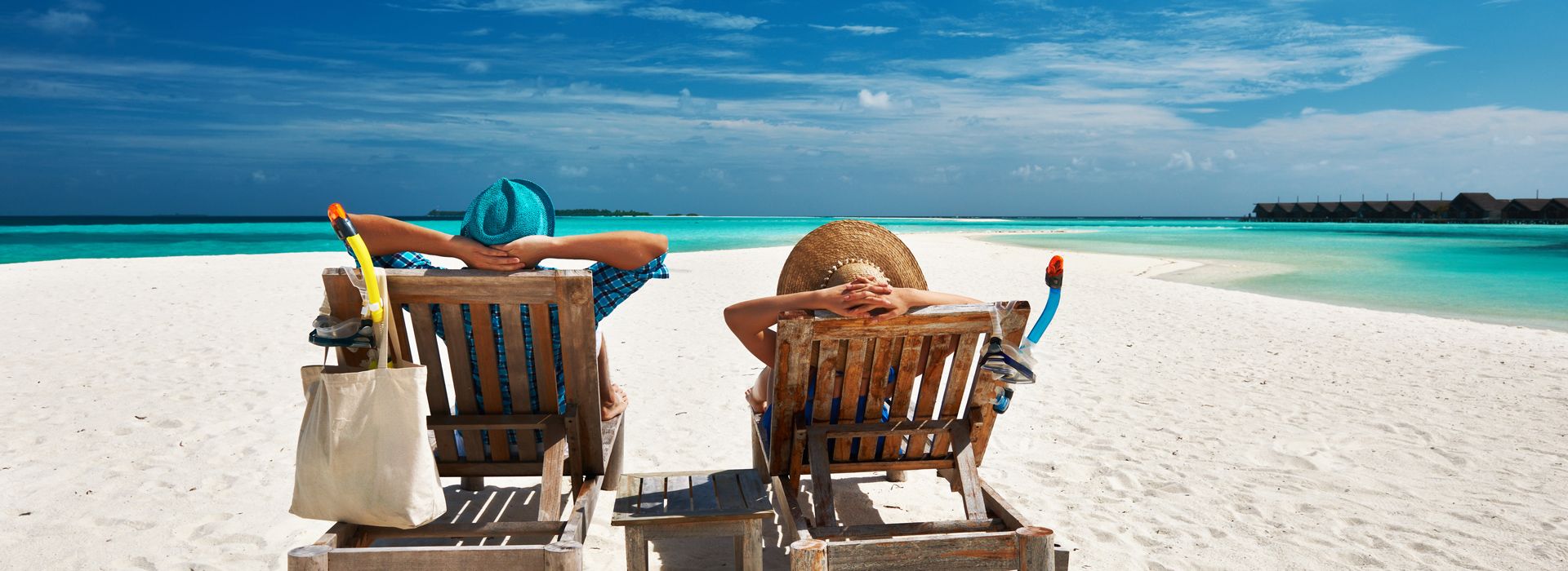 Mann und Frau mit Sonnenhüten liegen enspannt mit einer Auslandskrankenversicherung am Strand, türkisblaues Meer