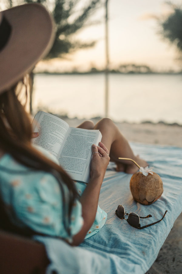 Frau mit Sonnenhut liegt am Strand und liest ein Buch, Kokosnuss-Getränk