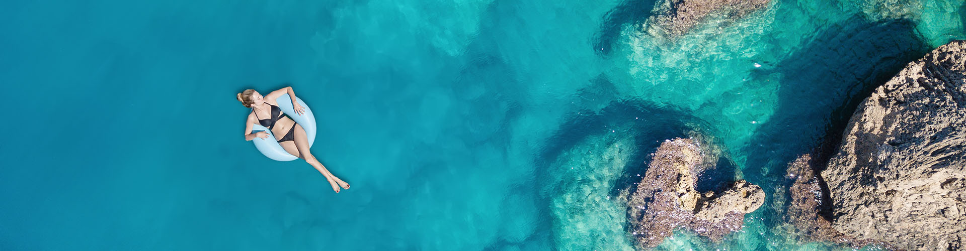 Frau auf einem Schwimmreifen schwimmend im türkisblauen Meer, felsige Küste