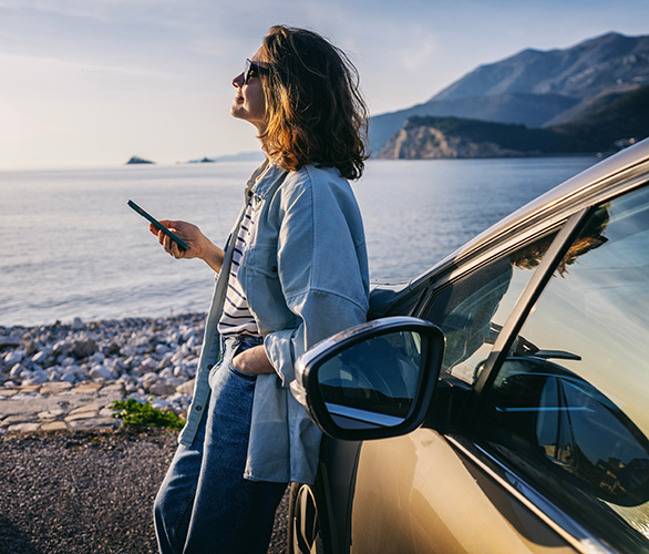 Lächelnde junge Frau mit Sonnenbrille an der Küste, angelehnt an ihrem Auto und mit dem Handy in der Hand