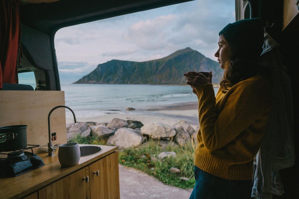 Frau lehnt an der Wand eines Campers und trinkt aus einem Becher, genießt Blick aufs Meer